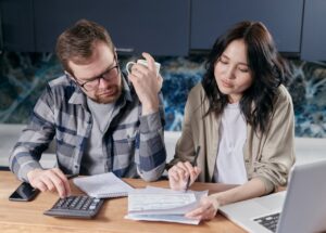 A woman and a man looking at the papers with the calculator and laptop on the desk.